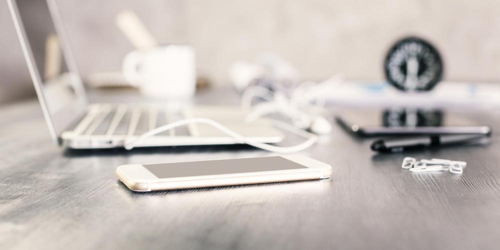 Weißes iPhone auf grauem Tisch mit Laptop und Tablet im Hintergrund 