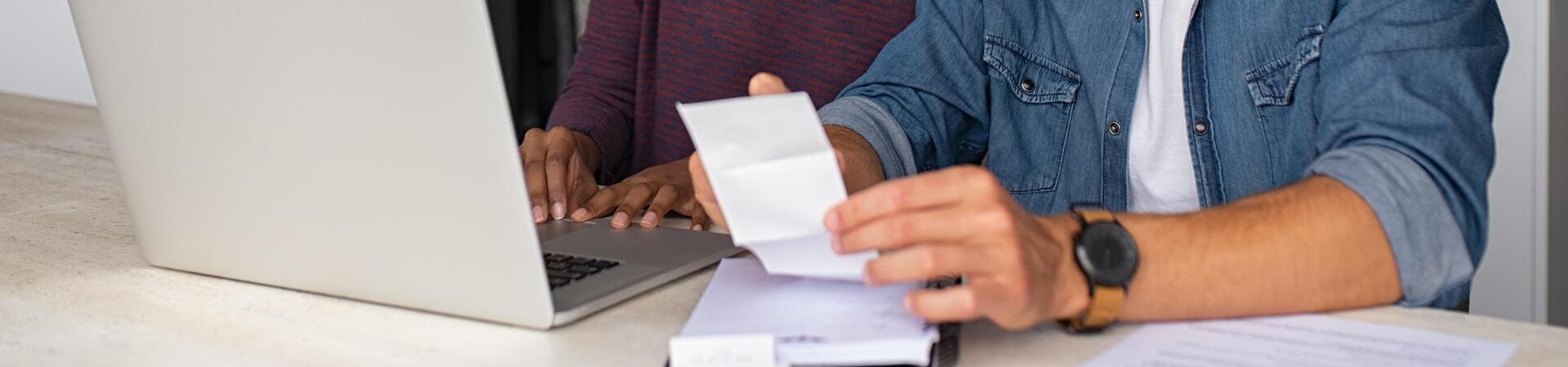 Zwei Menschen sitzen am Küchentisch vor einem Laptop und überprüfen gemeinsam Belege