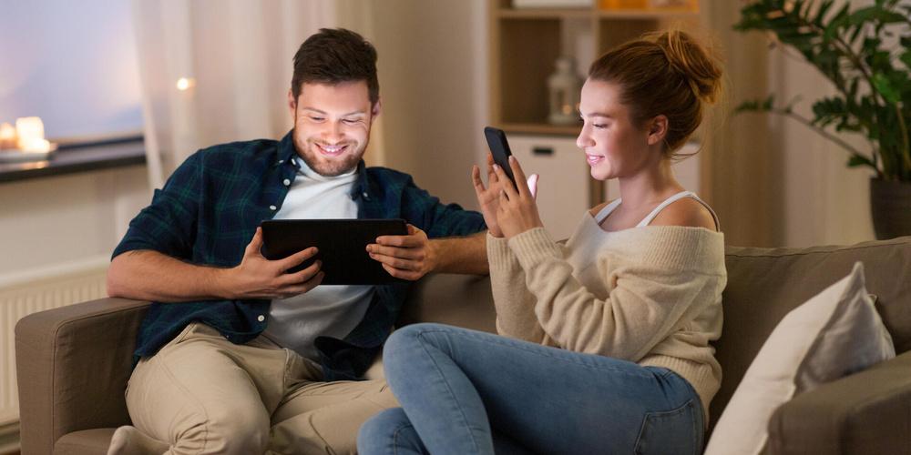 Zwei Personen nutzen Tablet und Smartphone gemeinsam auf dem Sofa